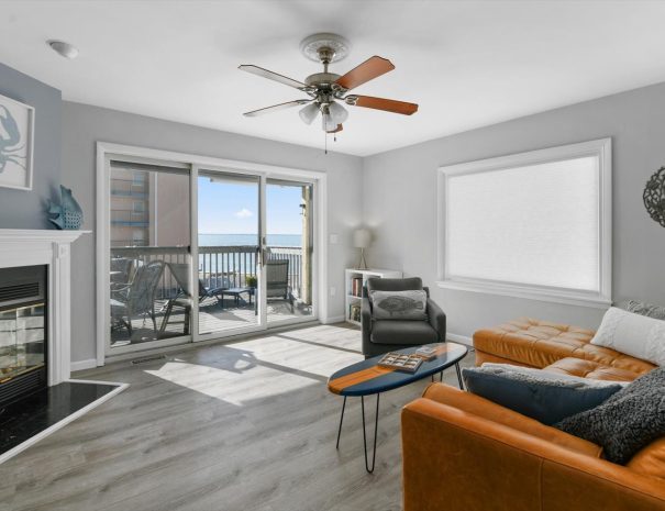 A modern living room with large sliding doors opening to a balcony with ocean view. It features a ceiling fan, tan leather sectional sofa, fireplace, gray armchair, round wooden coffee table, and wall art. The floors are light wood, and the walls are painted gray.