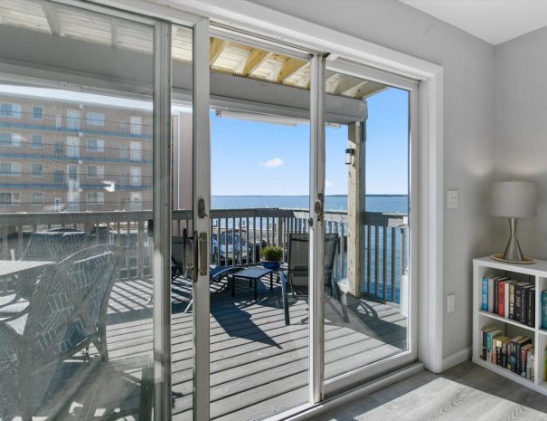 A bright room with a large glass sliding door leading to a wooden balcony overlooking the water. The balcony has outdoor furniture including a table, chairs, and a lounge chair. Inside, there's a bookshelf filled with books and a small lamp on top.