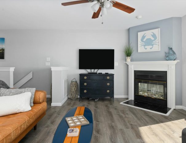 A modern living room with light gray walls, featuring a brown leather couch, a black chest with a flat-screen TV on top, a fireplace with a crab painting above it, and a ceiling fan. A surfboard-shaped coffee table sits on a wood floor.