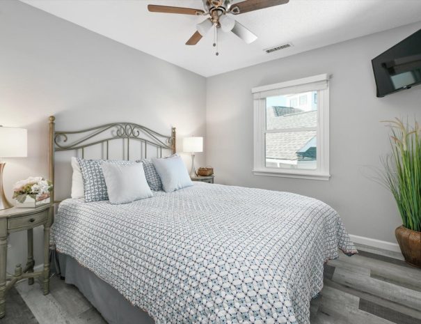 A cozy bedroom with a queen-sized bed featuring a patterned duvet and several pillows. The room has grey walls, wooden flooring, and a ceiling fan. There's a nightstand with a lamp and flowers on one side of the bed, and a flat-screen TV mounted on the wall.