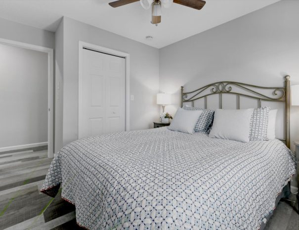 A bedroom with grey walls featuring a bed with a patterned blue and white duvet and pillows, flanked by two nightstands with lamps. A ceiling fan is above, and an open door and a closed closet are visible in the background. The floor is grey wood.