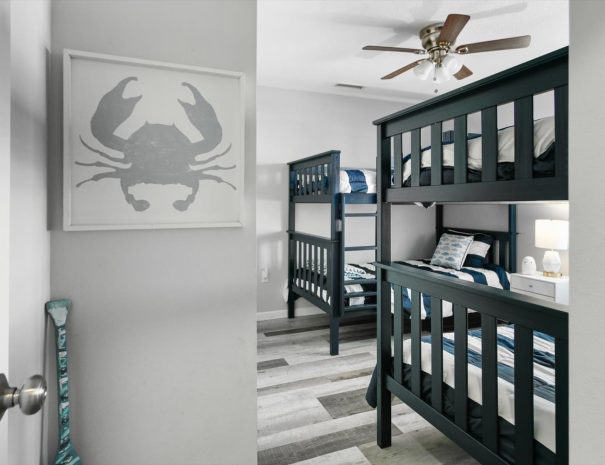 A bedroom with two dark green bunk beds, each adorned with striped bedding. The room has a light grey color scheme, wooden plank flooring, a ceiling fan, and a framed picture of a crab on the wall. A decorative paddle leans against the wall near the door.