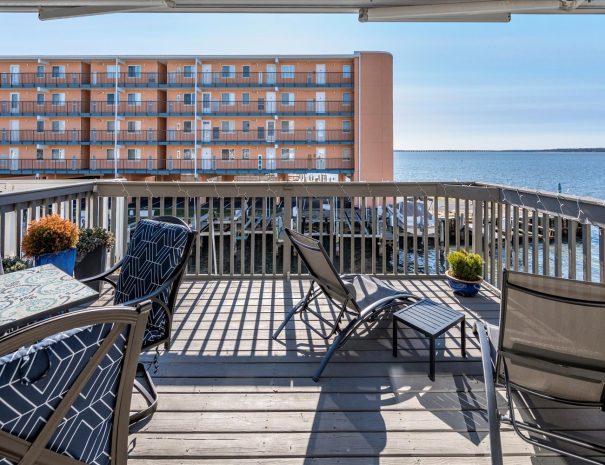 An outdoor deck with a table and patterned chairs, along with a few lounging chairs, overlooks a marina and water. In the background, there is an orange multi-story building with balconies. The deck is surrounded by a wooden railing and has planter pots with plants.