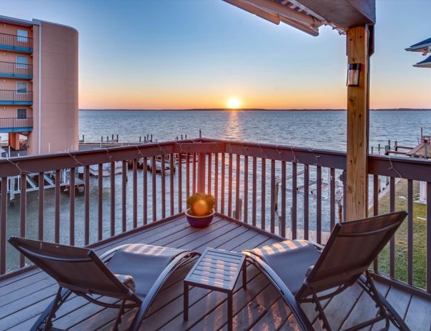 A balcony overlooking a calm body of water at sunset. Two lounge chairs face the setting sun, and a small potted plant sits between them. Nearby buildings and docks frame the water scene, creating a serene, picturesque environment.