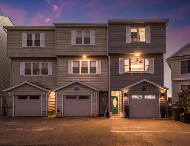 A three-story townhouse with a green front door, illuminated by exterior lights. The house has a garage on the ground floor, white-framed windows, and is flanked by neighboring homes. The twilight sky has shades of purple and pink, creating a warm ambiance.